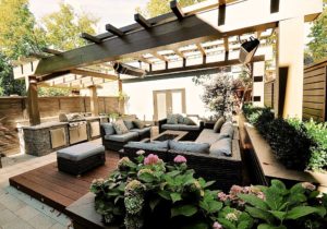 alt=raised planters wrap around a ground level deck that has an arrangement of outdoor furniture centred on a fire table | a pergola overtop spans the full width of the backyard with outdoor heaters suspended above the deck and a roof over an outdoor kitchen