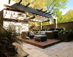 alt=deck podium clad in tropical hardwood | outdoor furniture arranged around a fire table | suspended has heaters | ornamental grass in foreground planter