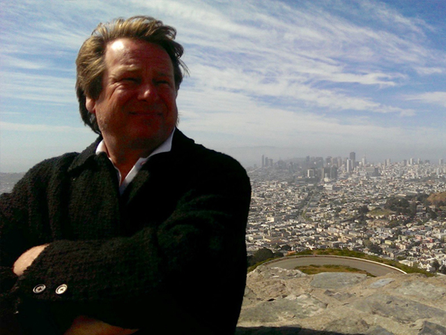 Photo of man with arms crossed, his face in shadow faces camera standing on a hill with downtown San Francisco in the background.