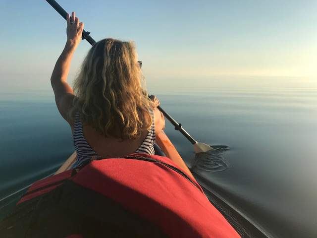 Looking from behind, a woman with long blond hair dips her kayak oar into glassy still water that continues to the horizon.